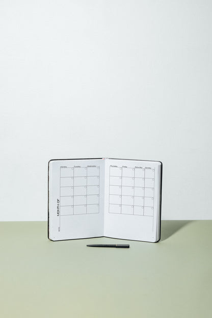 MSTRPLN Undated Planner in black, on a tan table with a green table with a black pen in front. Planner is opened to the undated monthly layout