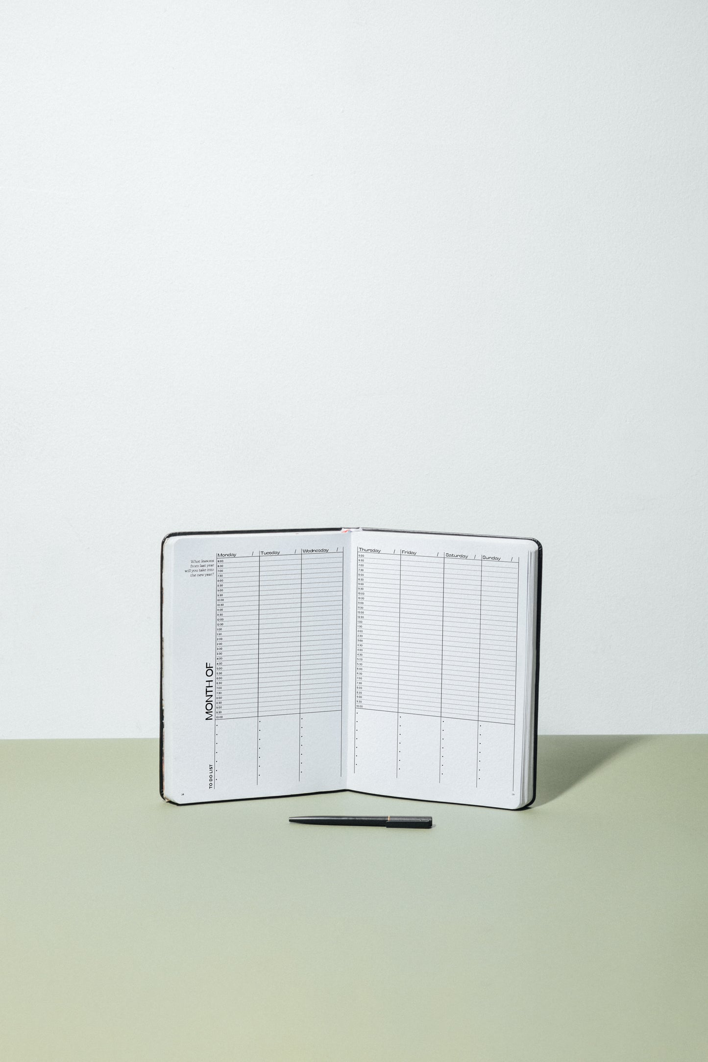 MSTRPLN Undated Planner in black, on a tan table with a green table with a black pen in front. Planner is opened to the undated weekly layout